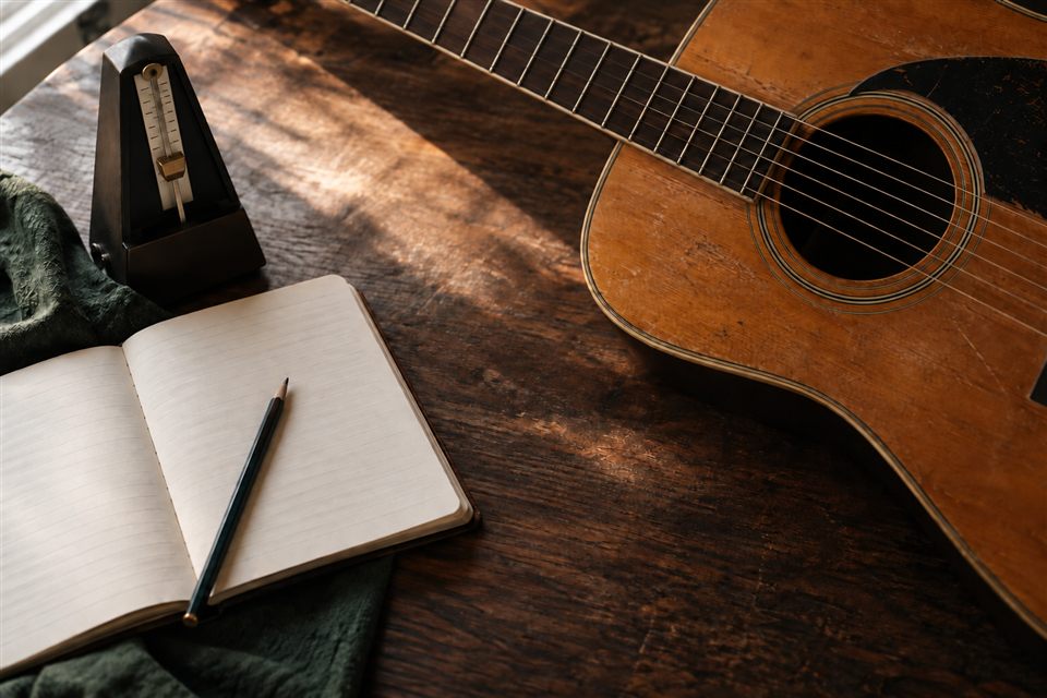 A worn acoustic guitar, metronome and open practice notebook on a wooden table.